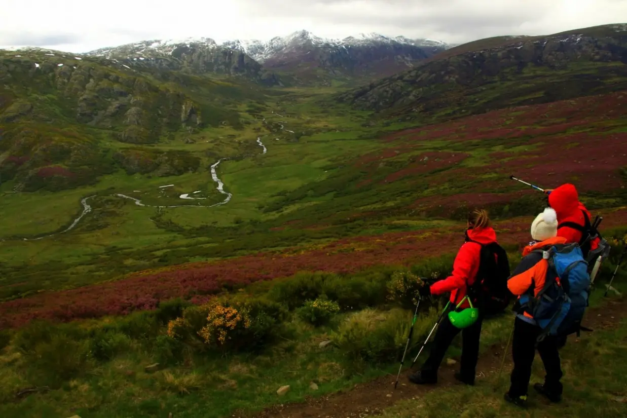 Dos senderistas contemplando un valle verde con cumbres nevadas al fondo